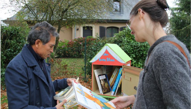 une bibliothécaire réalimente une boite de dons de livres tandis qu'un citoyen consulte un ouvrage déposé