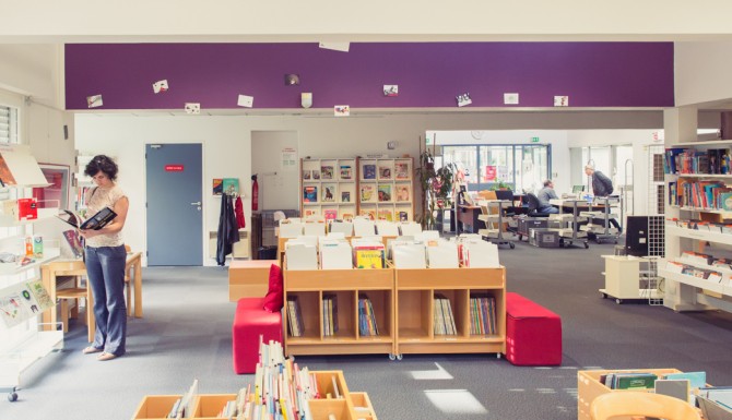plan large de la bibliothèque, mur du fonds violet, on y voit les espaces avec leurs mobiliers et des usagers. Au fonds, on aperçois l'accueil