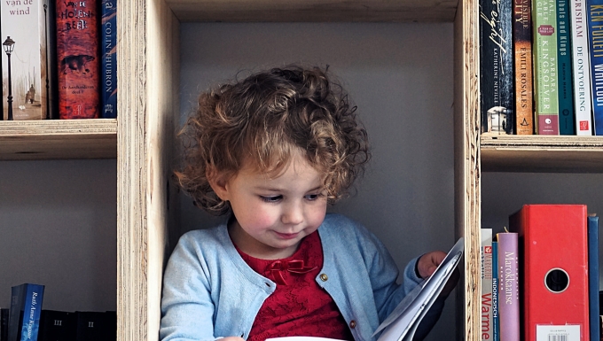 Une petite fille lit un livre assise dans le bas d'une bibliothèque en bois.