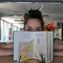Jeune femme avec la partie basse du visage dissimulée derrière un livre. Paire de lunettes au premier plan.